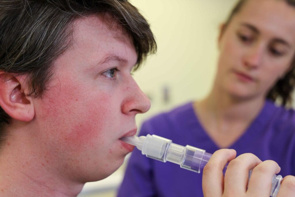 A man breathing with a secretion clearance device with a nurse in the background